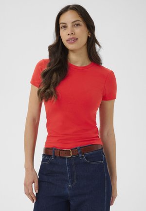 Femme aux longs cheveux bruns portant un t-shirt rouge ajusté à manches courtes, un jean bleu foncé et une ceinture en cuir marron, debout devant un fond uni.