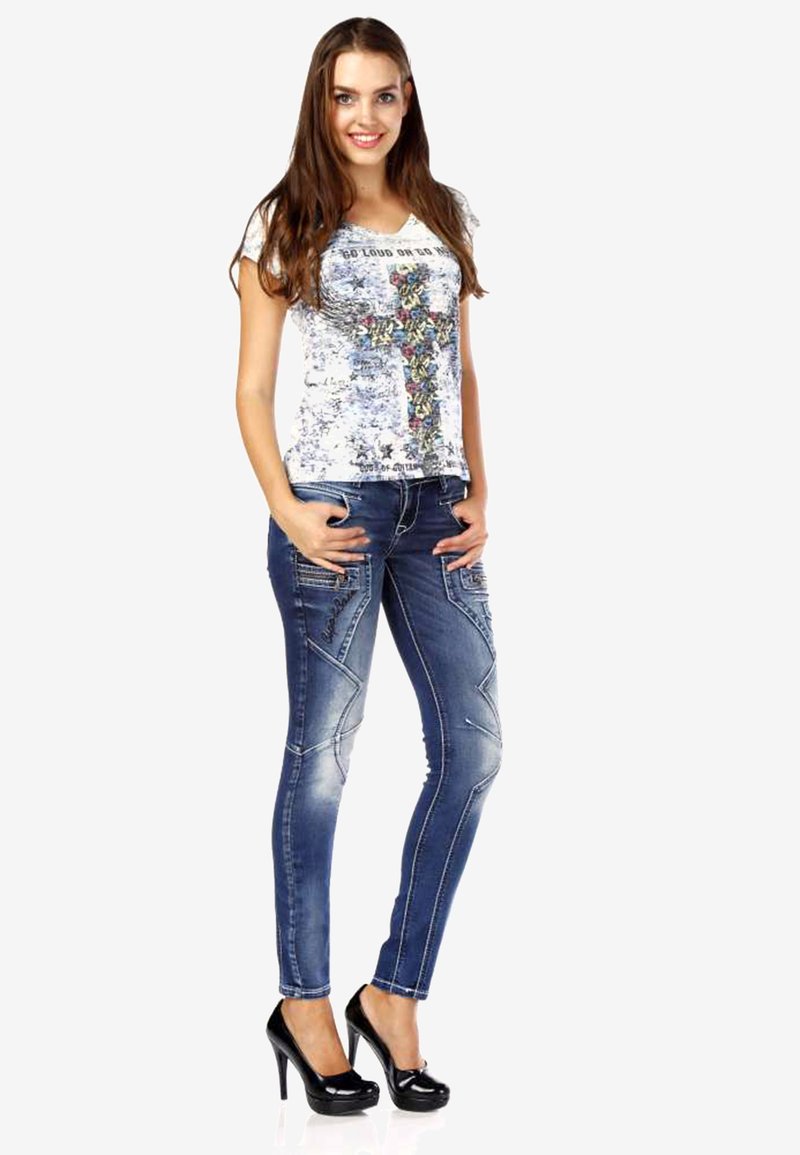Blaue, hautenge Denim-Jeans mit diagonalen Nahtdetails, kombiniert mit einem weißen T-Shirt mit einem bunten Grafikdesign und Text. Schwarze High Heels.