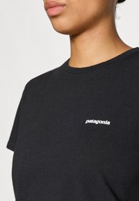 Melna kokvilnas T-kreklu ar apaļu kakla izgriezumu un īsām piedurknēm, uz kreisā krūšu laukuma ir balts "Patagonia" logo.