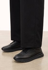 Sneakers din piele neagră cu un finisaj texturat, având vârf rotund, design cu șireturi și o talpă din cauciuc texturată. Purtați cu pantaloni negri.