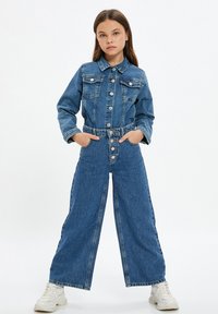 Meisje in een knoopje dichtgenaaide denim jas en hoog- taille wijde jeans met handen in de zakken, staand met gespreide benen in witte sneakers.
