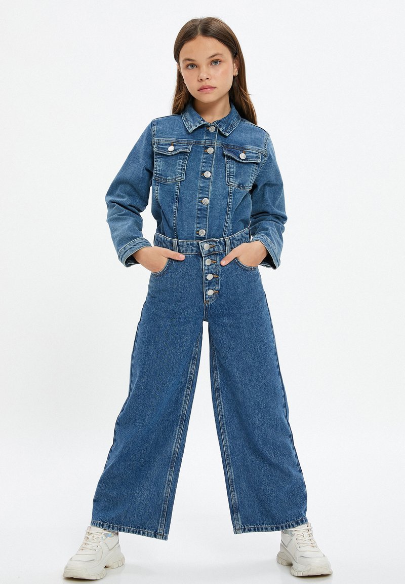 Meisje in een knoopje dichtgenaaide denim jas en hoog- taille wijde jeans met handen in de zakken, staand met gespreide benen in witte sneakers.