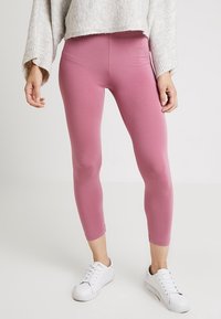 Person klädd i korta rosa leggings, vita sneakers och en ljusgrå stickad långärmad tröja, stående mot en enfärgad bakgrund.