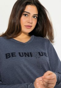 Dunkelblaues Sweatshirt mit V-Ausschnitt, auf dem der Satz "SEI EINZIGARTIG" in kontrastierendem Schwarz gestickt ist. Der Stoff wirkt weich und texturiert.