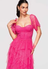 Femme en robe en dentelle rose vif avec jupe en tulle superposée et manches courtes en dentelle, posant la main près de l'épaule et regardant sur le côté.