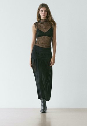 Schwarzes transparentes Oberteil mit strukturierter Detailverarbeitung, kombiniert mit einem figurbetonten schwarzen Rock mit Falten und einer gewellten Taille. Schwarze kniehohe Stiefel vervollständigen den Look.