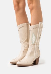 Botas de cuero crema hasta la rodilla con diseño ondulado, acentos decorativos en forma de hoja, cremallera plateada y tacón de bloque de madera. Acabado texturizado.