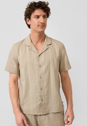 Homme aux cheveux bouclés portant une chemise beige à manches courtes boutonnée et un pantalon assorti sur un fond clair uni.