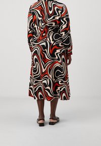 Vestido de manga larga con un patrón en espiral en negro, rojo y beige, con cintura elástica, que llega a medio pantorrilla, combinado con sandalias planas.