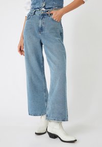 Jeans de mezclilla anchos de color azul claro con diseño clásico de cinco bolsillos, cierre de botón metálico y dobladillo deshilachado. Combinados con botas de cowboy blancas.