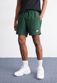 Mörkgröna Nike-shorts med elastiskt midjeband och sidofickor, med en vit Nike-logotyp nära fållen. Bärs med vita sneakers och strumpor.