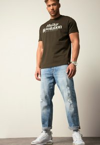 Camiseta de algodón verde oliva con texto blanco. Jeans de mezclilla desgastados en azul claro. Zapatillas blancas con detalles grises. Reloj plateado en la muñeca.