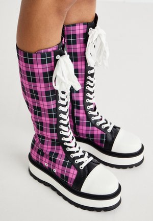 Bottes à plateforme montant jusqu'aux genoux avec un motif à carreaux rose et noir, bouts blancs, bordures noires et lacets blancs portées sur des jambes nues.