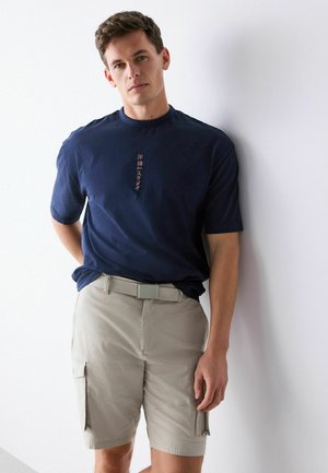 Mann lehnt an einer weißen Wand, trägt ein marineblaues T-Shirt mit vertikalem roten Text und beige Cargo-Shorts mit Gürtel.