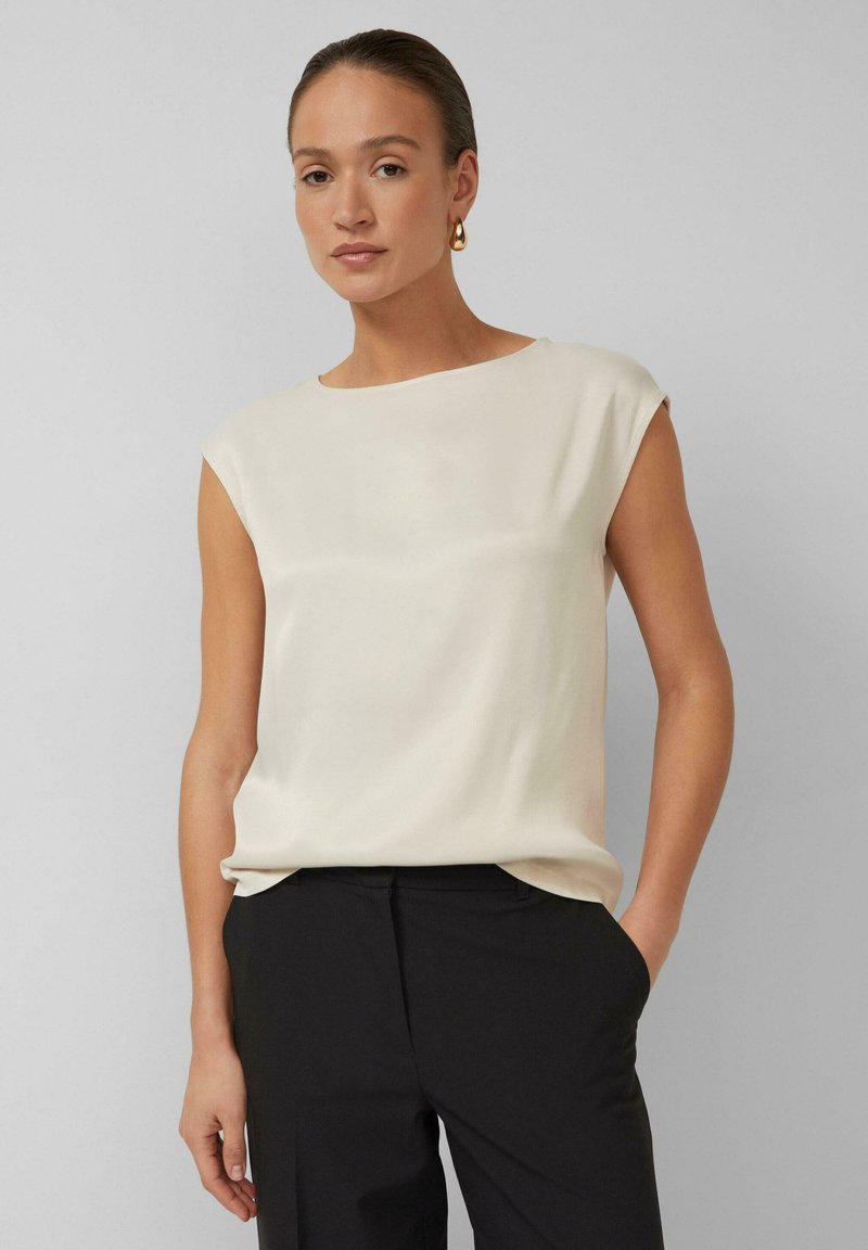 Vrouw met naar achteren gestylede haren draagt een mouwloze crèmekleurige blouse, zwarte broek en gouden oorringen, staand met één hand in de zak.