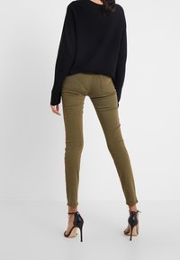 Pantalones ajustados verde oliva con bolsillos traseros, combinados con un suéter negro de corte oversize y sandalias de tacón negro.