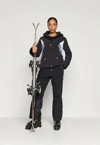 Σακάκι σκι σε μαύρο χρώμα με ανοιχτό μπλε λεπτομέρειες και κουκούλα, σε συνδυασμό με μαύρα παντελόνια και μπότες σκι, κρατώντας σκι και μπαστούνια.