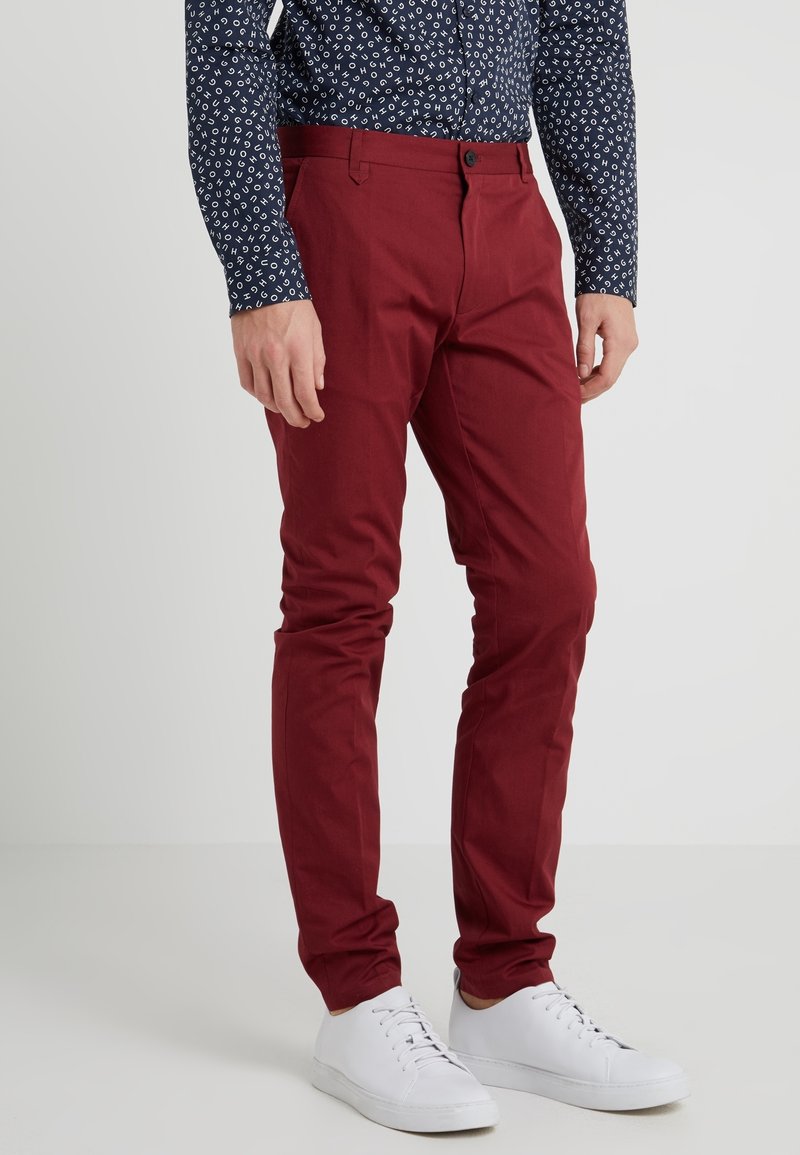Homme portant un pantalon slim rouge foncé, une chemise bleu marine à motifs et des baskets blanches, debout devant un fond uni.