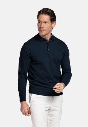 Man die een zwart poloshirt met lange mouwen en knopen draagt en een witte broek, staand en zijwaarts kijkend tegen een effen achtergrond.