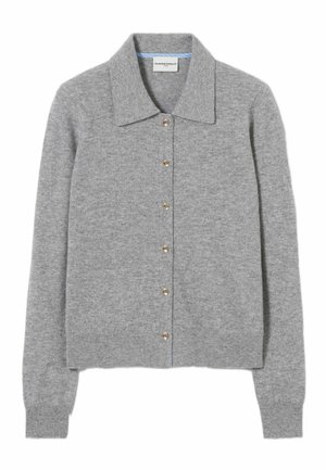 Femme portant un cardigan gris boutonné sur une robe bleu marine, ceinture noire, tenant un petit sac à main bordeaux et des sandales dorées à brides, debout.