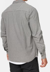 Man die een grijze denim overhemd met lange mouwen over een wit T-shirt en een zwarte broek draagt, vanachter gezien met een ontspannen armhouding.