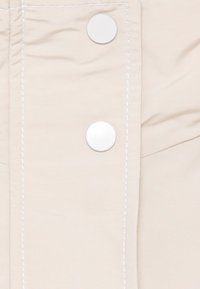 Tecido bege com costura branca visível e dois grandes botões de pressão brancos, apresentando uma textura suave e um design simples.