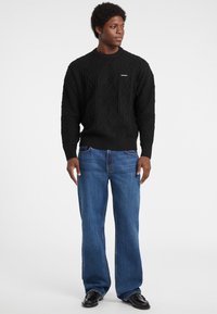Maglione nero lavorato a maglia con un motivo a diamante, maniche e orlo a coste. Abbinato a jeans a gamba larga blu e scarpe nere.