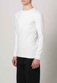 T-shirt branco de manga longa com decote redondo, feito em tecido de algodão suave, de forma ajustada e com detalhes sutis de costura nos punhos.