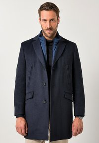 Granatowy wełniany płaszcz z fakturowanej tkaniny, z klapami z nacięciem, dwoma przednimi kieszeniami oraz ukrytym zamkiem pod kołnierzem.