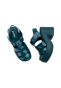 Aksamitowe sandały na platformie w odcieniu teal, wykonane z błyszczącego, elastycznego materiału, z otwartym noskiem, wieloma paskami oraz grubym obcasem. Złote akcenty na klamrze.