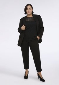 Žena stojí sebevědomě v černém saku, vzorovaném černém topu, černých kalhotách a černých podpatcích na jednobarevném světlém pozadí.