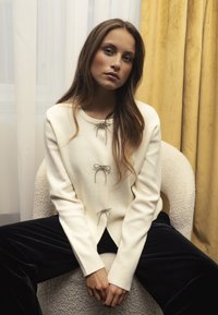 Cream-colored cardigan made of soft material, featuring three silver bow accents and a round neckline, paired with dark velvet pants.