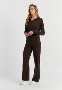 Bruin gebreid lounge-outfit bestaande uit een top met kraag en wijde broekspijpen. Zachte textuur met subtiele details. Draagt met witte sneakers.