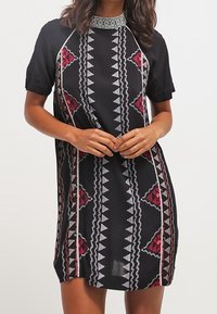 En kvinna poserar i en kortärmad svart klänning med röda och vita geometriska mönster, stående med händerna knäppta framför sig.