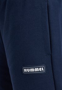 Zbliżenie na granatową tkaninę z bocznym szwem i prostokątną naszywką z logo HUMMEL w pobliżu brzegu.