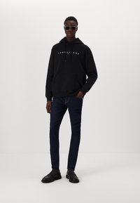 Svart bomullshoodie med frontlomme og hvit "TOMMY JEANS" logo, sammen med mørkeblå jeans og svarte sko. Modell med solbriller.