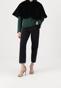 Svart cape i konstpäls över en grön ribbad tröja. Modellen bär mörka korta jeans och beige remsandaletter med klack. Enkel design utan mönster.