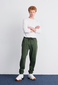 Pantalones deportivos verdes con cintura elástica y cordón, combinados con una camiseta de manga larga blanca y zapatillas blancas con detalles naranjas.