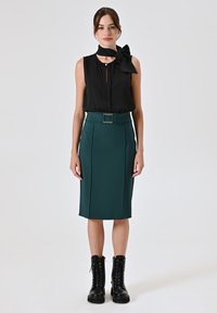 Blusa nera senza maniche con cravatta al collo, abbinata a una gonna a matita aderente color te, dotata di una cintura. Il modello indossa stivali neri con lacci.