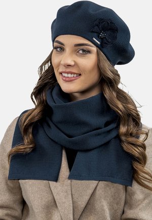 Marineblauer gestrickter Barett mit einer strukturierten Blumenakzentuierung und passendem Schal, beide aus einem weichen Stoff mit sauberen Kanten. Getragen mit einem hellbraunen Mantel.
