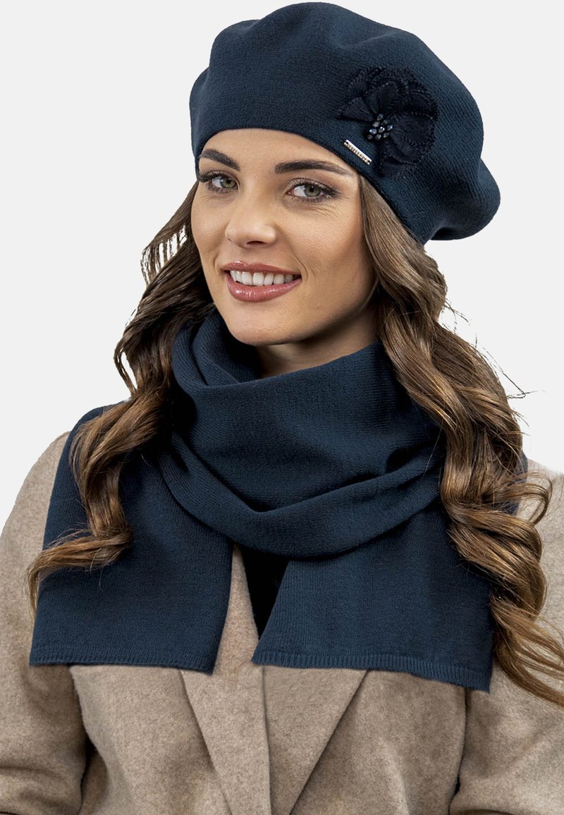 Marineblauer gestrickter Barett mit einer strukturierten Blumenakzentuierung und passendem Schal, beide aus einem weichen Stoff mit sauberen Kanten. Getragen mit einem hellbraunen Mantel.