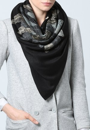 Femme portant un foulard triangulaire noir avec des motifs de feuilles gris et beige sur un manteau croisé gris clair, une main dans la poche.