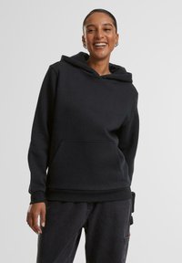 Persoană zâmbitoare purtând un hanorac negru cu buzunar frontal și pantaloni cargo negri, stând în fața unui fundal simplu și deschis la culoare.