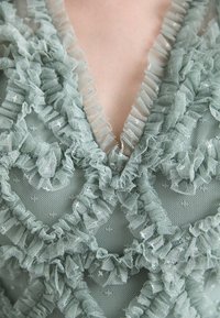 Tissu vert menthe orné de volants superposés, doté d'un col en V et d'un motif texturé délicat. Doux, transparent et détaillé.