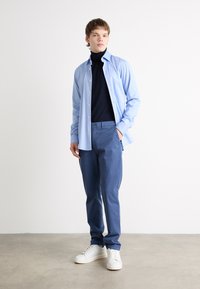 Lyseblå knapp-up skjorte over en svart turtleneck, kombinert med blå bukser og hvite joggesko. Glatt stoff, slim fit design.