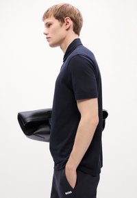Joven vestido con polo negro y pantalones, sosteniendo un bolso de cuero negro plegado bajo su brazo, de pie en perfil frente a un fondo blanco.