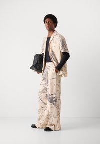 Beige fabric outfit with map print, short-sleeved shirt and wide-leg pants. Black long-sleeve top underneath, holding a black handbag.