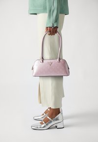 Borsa a tracolla rosa con due manici, chiusura a zip e logo in evidenza. Il modello indossa scarpe col tacco argento e un top verde menta con pantaloni bianchi.