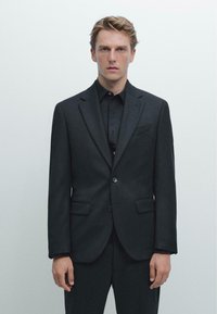 Un abito nero su misura con giacca a una fila di bottoni, rever a incasso e due tasche frontali. Indossato sopra una camicia nera con texture opaca.