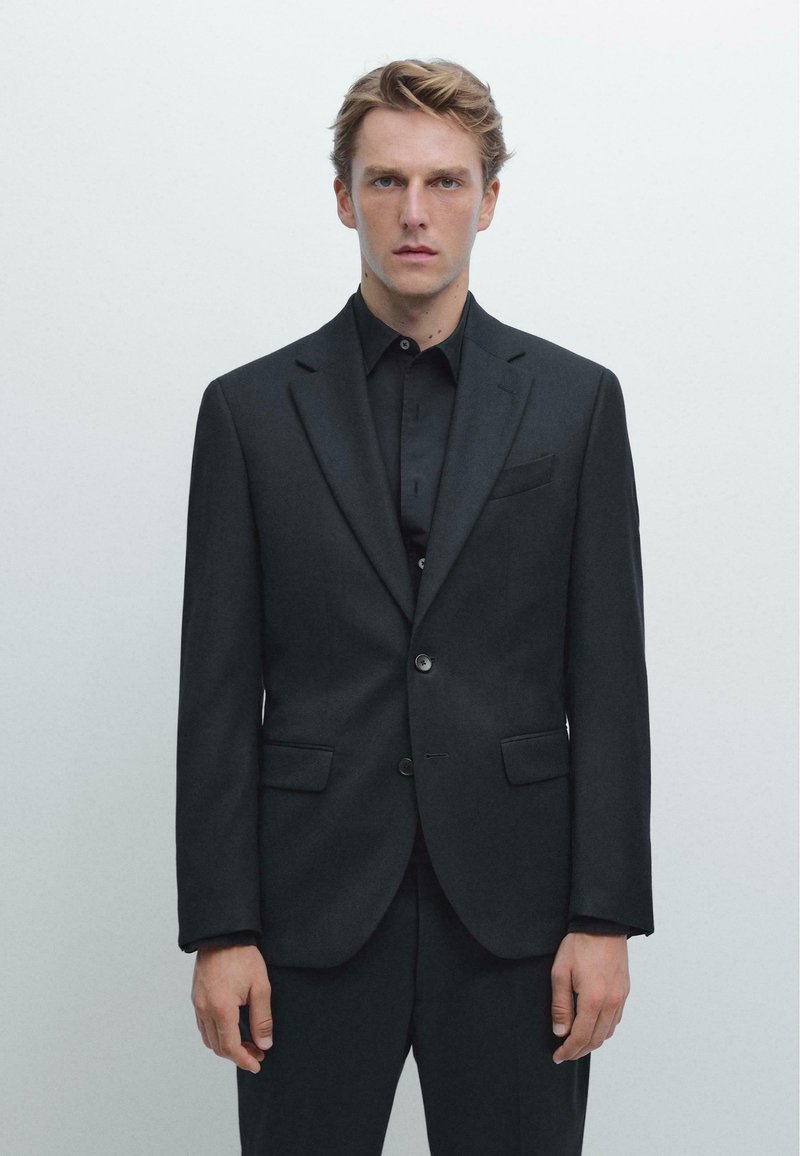 Un abito nero su misura con giacca a una fila di bottoni, rever a incasso e due tasche frontali. Indossato sopra una camicia nera con texture opaca.